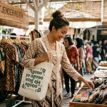 Foto dokumenter estetik seorang wanita Indonesia berusia akhir 30-an dengan sanggul rambut kasual dan kebaya kutu baru brokat krem sedang tersenyum di pasar kerajinan tangan Yogyakarta. Ia menyandang tote bag kanvas putih merek Tjenderakata yang menampilkan tipografi hand-lettering hijau bertuliskan "Ngono yo ngono ning aja ngono". Di depannya, ia sedang melihat-lihat produk pouch kecil dari kain perca sutra yang dipajang di stan pasar kayu dengan latar belakang yang agak blur.